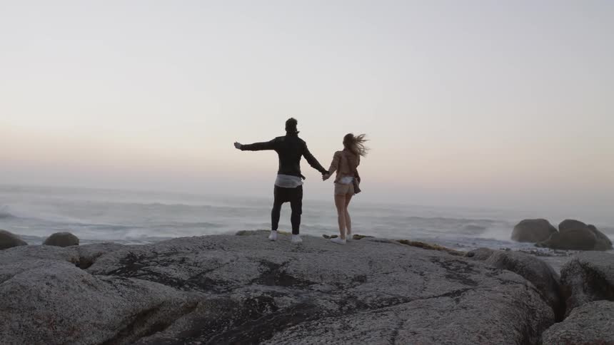 A man and woman stand on a rock above the ocean as the wind blows