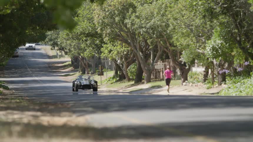 Black car drives down a tree lined country road past a woman jogging