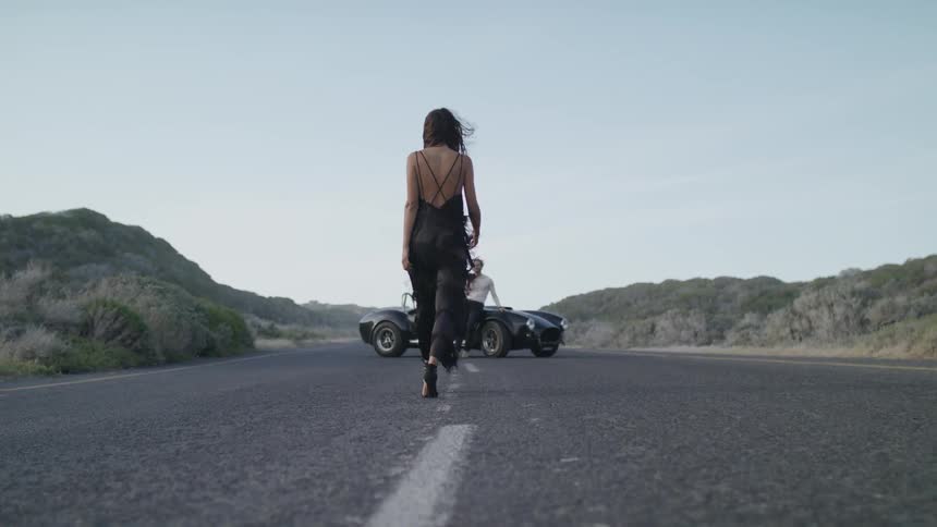 Woman in a dress walks towards a man leaning against a car