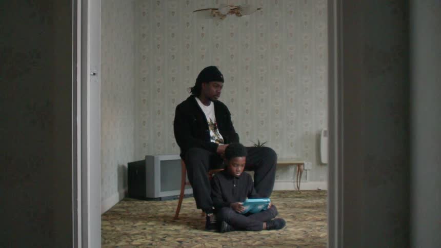 A man sits on a chair in a room while his son plays on the floor