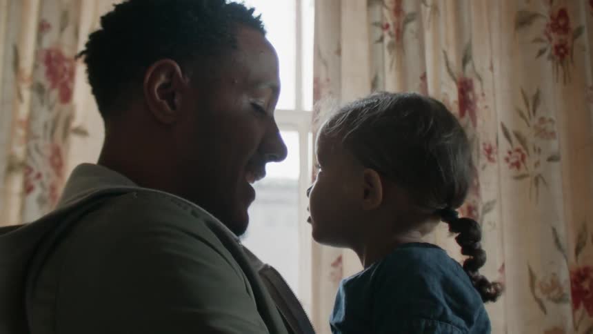 A man is giving his daughter a kiss as they sit inside in daylight
