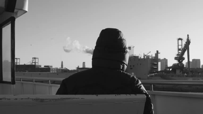 Someone wearing a hooded jacket sits on the ferry as it passes a factory