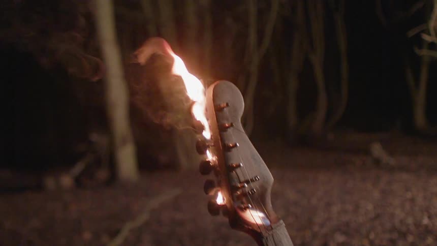 The head of a guitar burns in the woods at night