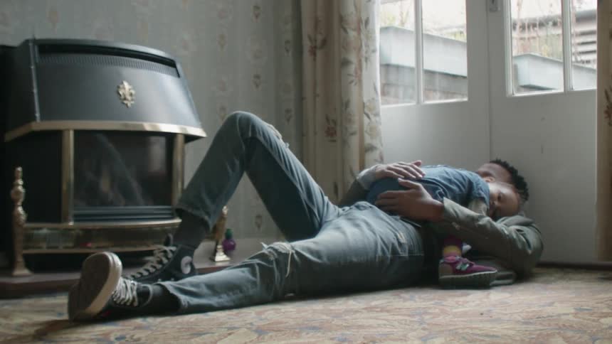 A man is patting the back of a sleeping toddler as they lie on the ground inside during the day