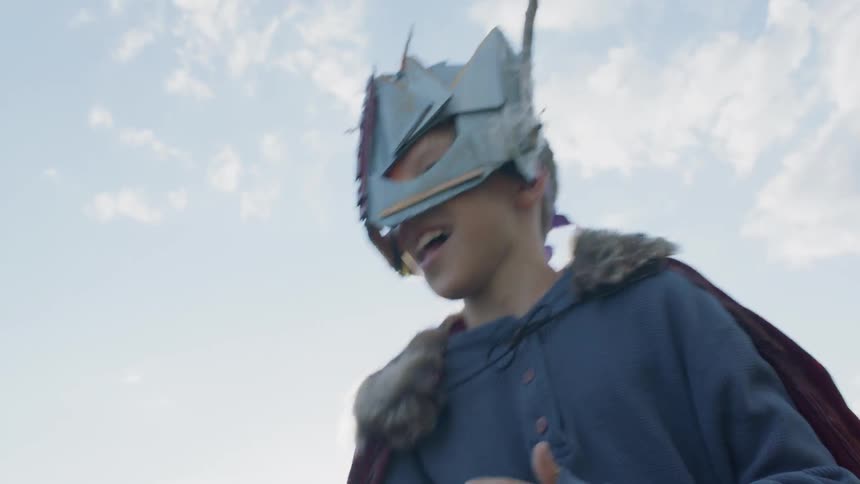 Young child wearing a cape and mask swings a stick around