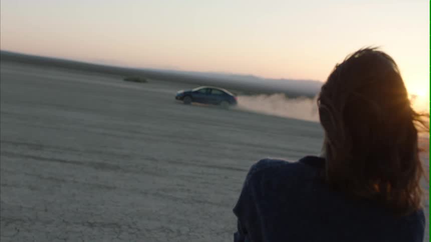 A woman stands and watches a car as it drives circles in the desert sand