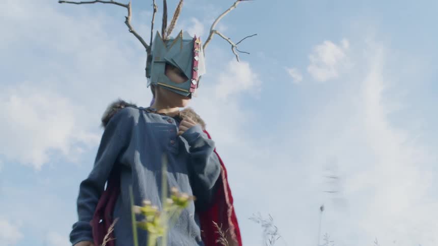 Child wearing a mask and a cape waves a stick hitting weeds