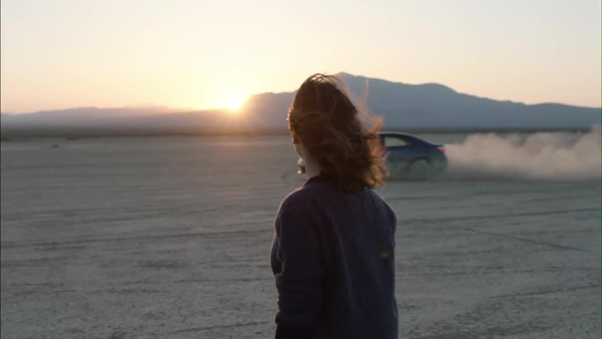 A woman walking through an open field at sunset as a car drives by
