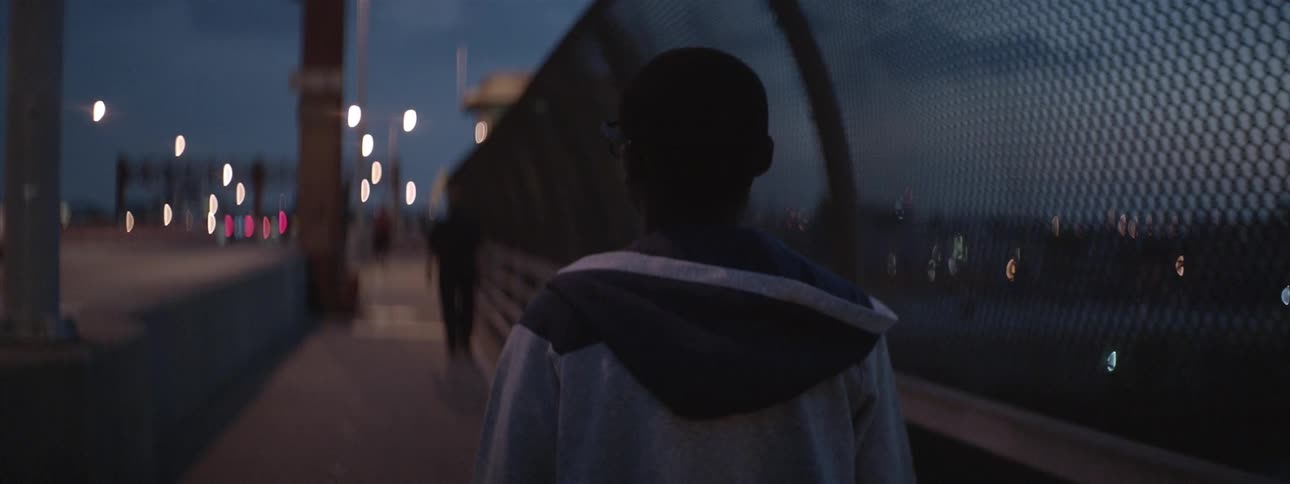 A boy walks along a bridge at night wearing a hoodie