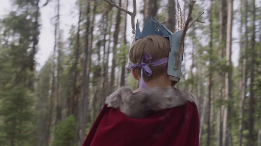 A boy wearing a mask and cloak is exploring a forest in daylight
