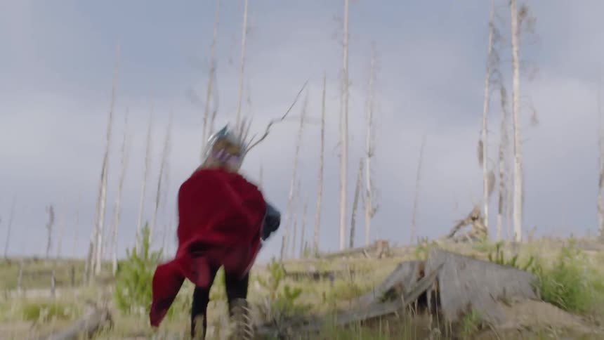 Child wearing a red cape and mask runs through a field with tree stumps