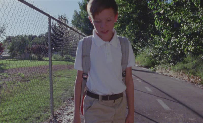 Child wearing a backpack beats a stick against a chain link fence