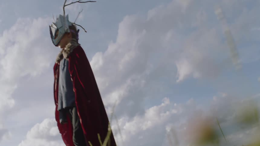 Young child wearing a cape and mask stands under the cloudy blue sky
