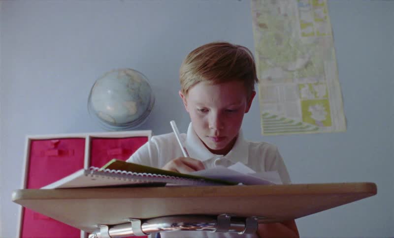 A boy is writing while sitting at a school desk inside in daylight