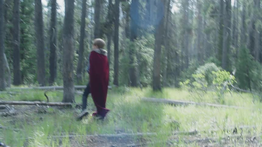 Boy wearing a cape runs through the forest as he throws things