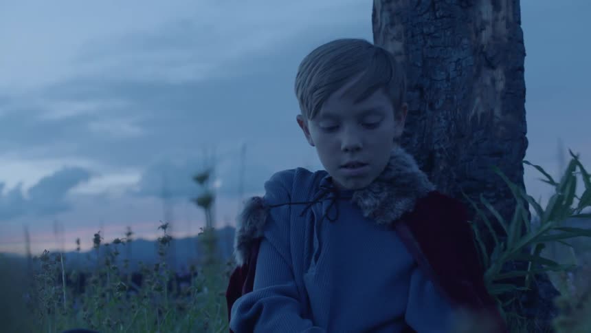A boy wearing a cape and holding a mask sits against a tree