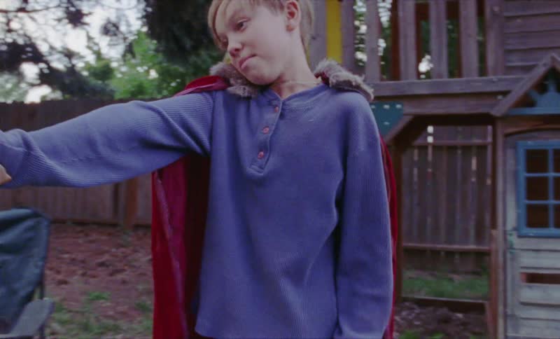 Young boy in a red cape being chased by two other children in a yard