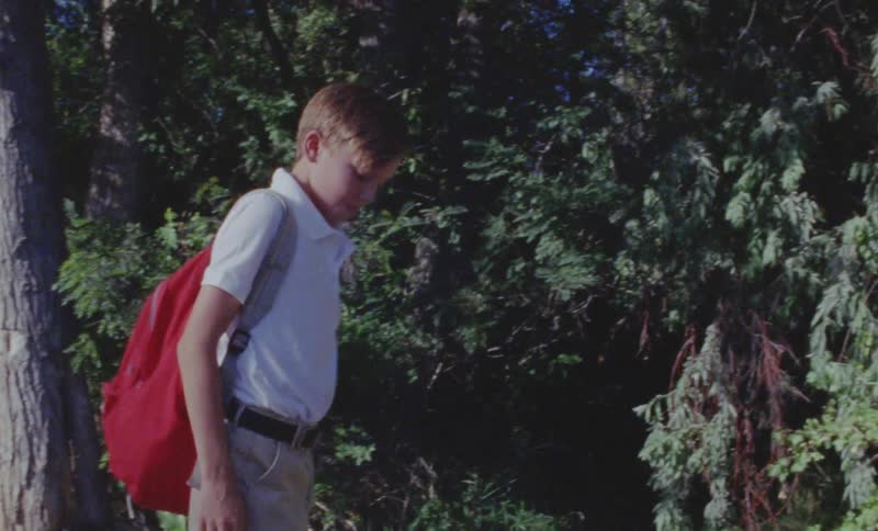 A boy drops his backpack at the edge of the tree line