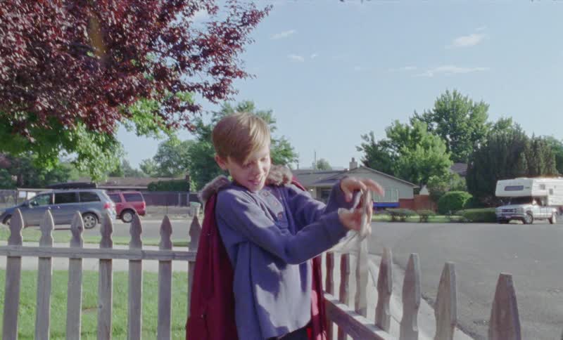 Child wearing a cape and carries a stick walking along a white picket fence