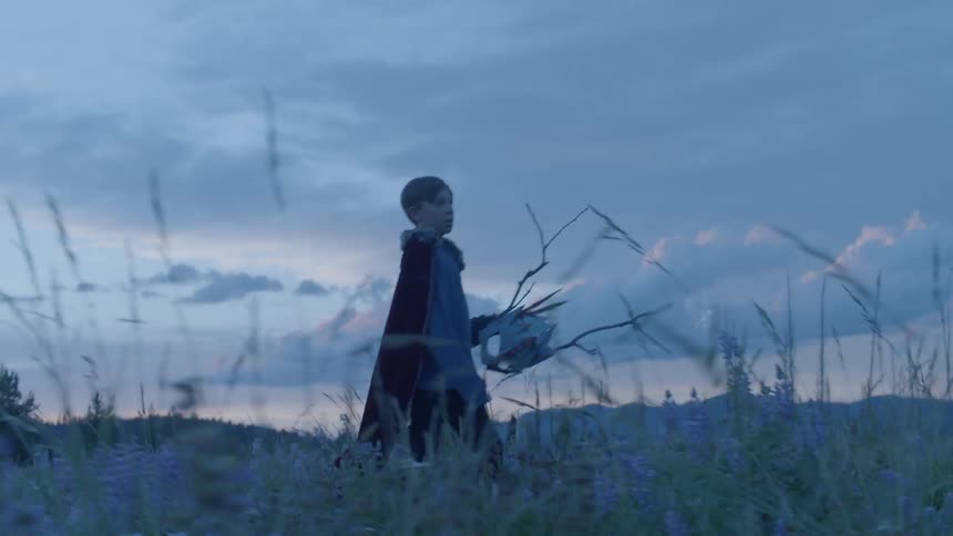 Young boy wears a red cape and carries a mask as he walks in a field