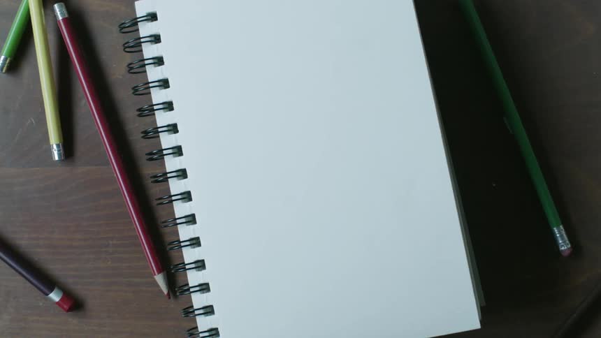 Pencils lie scattered near a blank notebook on a table in daylight