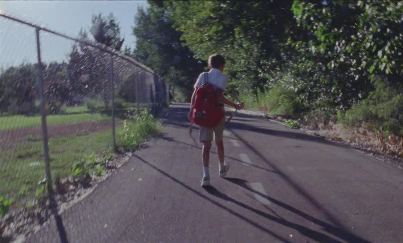 Boy with a red backpack walks down the road carrying a stick