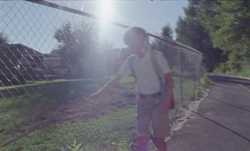 The child is walking past the fence on a sunny day