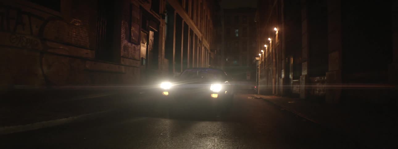 A car with headlights on moves down a narrow city road at night