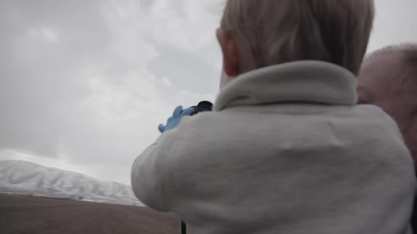 An old man is holding a toddler and helping him look through binoculars on a cloudy day
