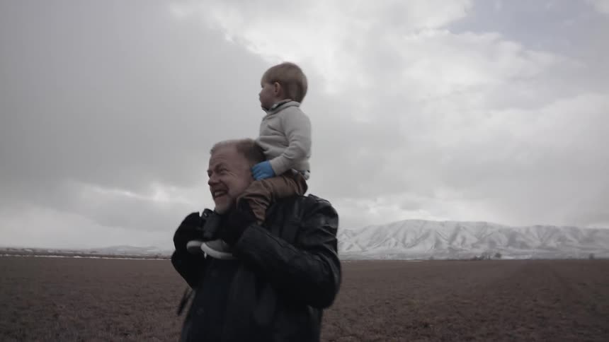 A grandpa is carrying his grandson on his shoulders in a natural environment on a cold day