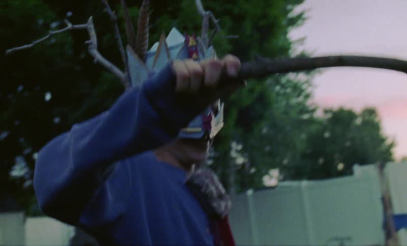 Child wearing a mask waves a stick in a yard at sunset