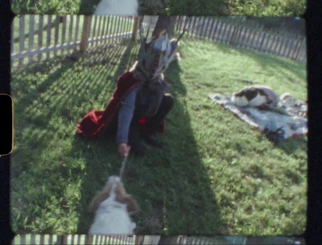 Young boy wearing a mask and red cape holds a stick as a dog pulls on it