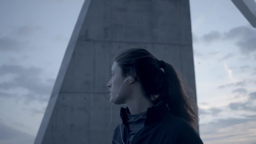 Woman wearing earbuds stands outside near a large structure at sunset