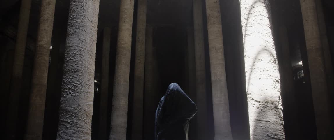 Person shrouded in a black cape moves in between many pillars