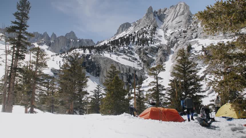 People camp near trees at the base of mountains in the snow