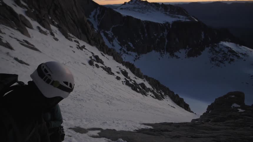 Person wearing a helmet stands on a snowy mountain top at sunset