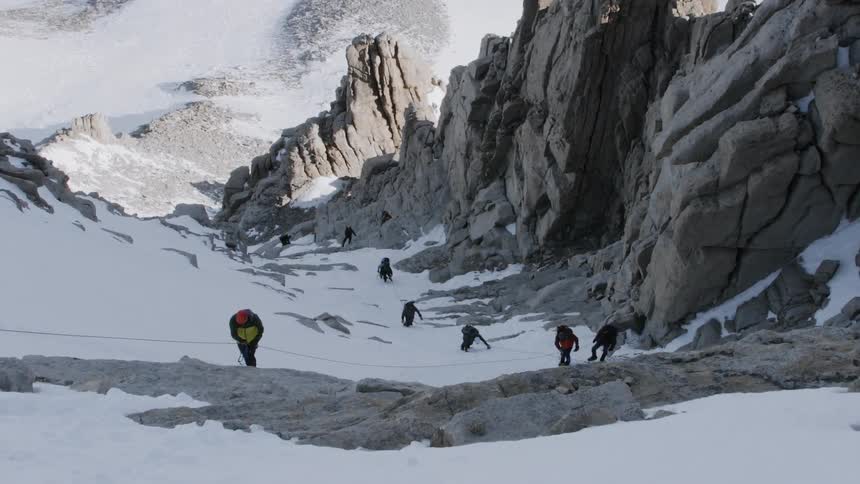 Many people hike up a snow covered mountain holding onto a rope
