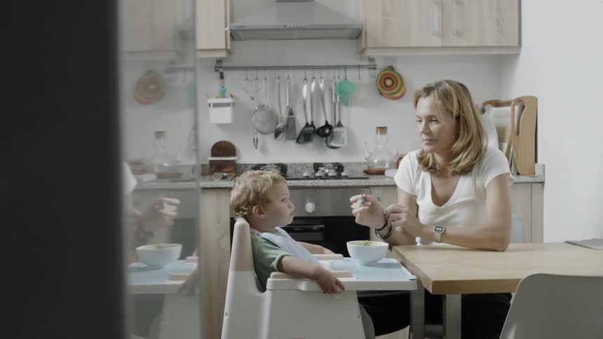 Woman feeds a baby as a man stands at the kitchen counter