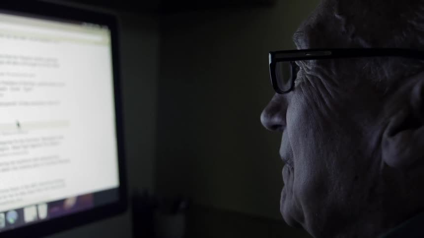 An older man is squinting as he looks at a computer screen in the dark