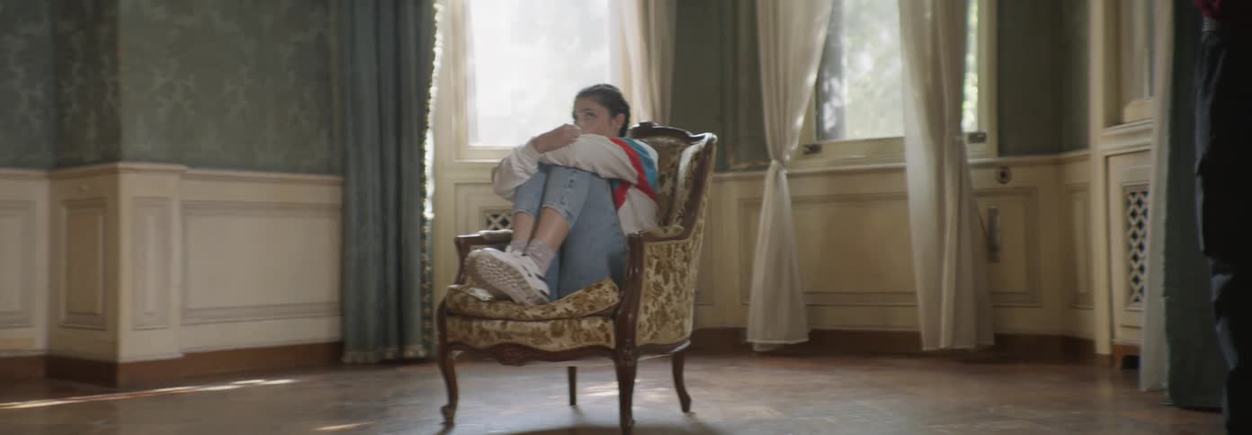 A girl is pulling her knees to her chest as she sits in a chair in a room in daylight
