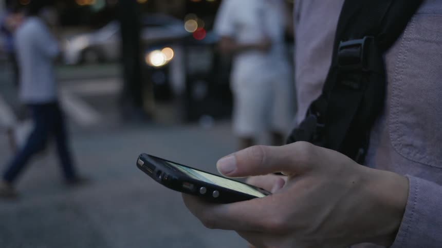 A man is standing outside on a sidewalk in a city at dusk using a cellphone