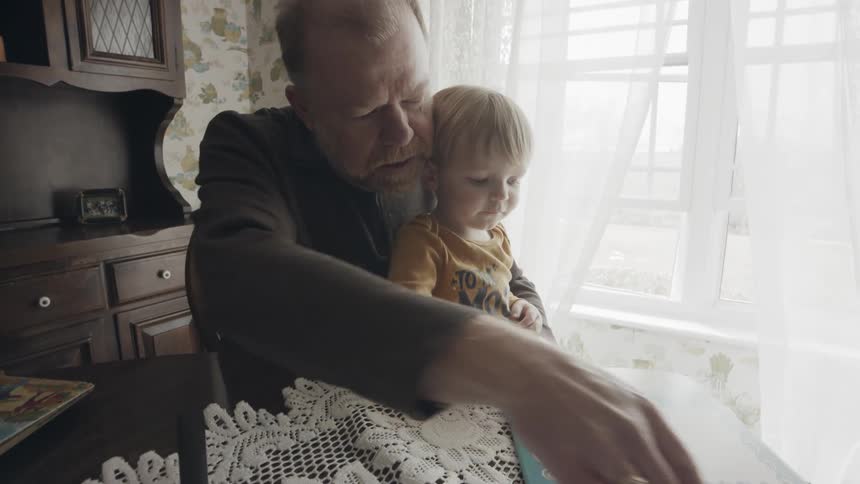 A grandpa is holding his grandson on his lap as they read at a kitchen table in daylight