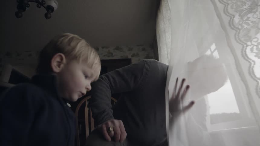 An old man is waving through a window curtain as he plays inside with a toddler in daylight