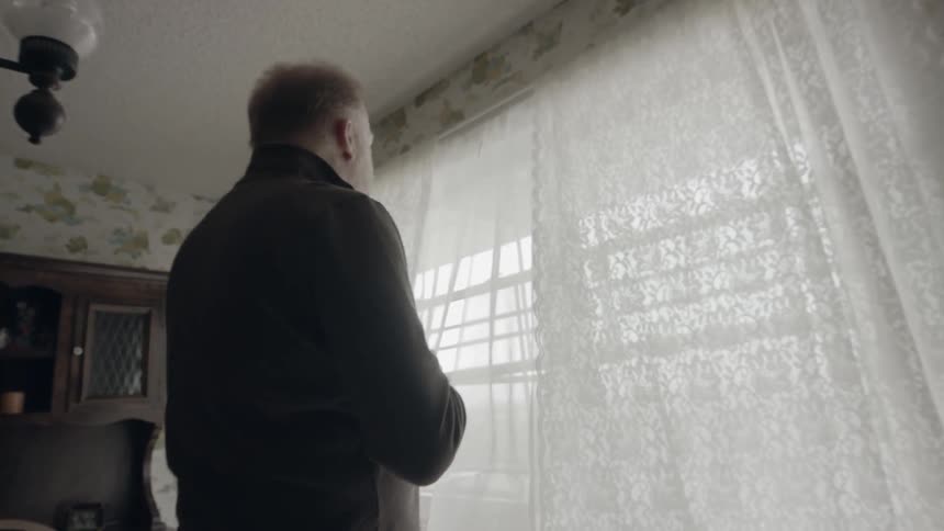 An old man is holding a coffee cup while he looks out of a window in daylight