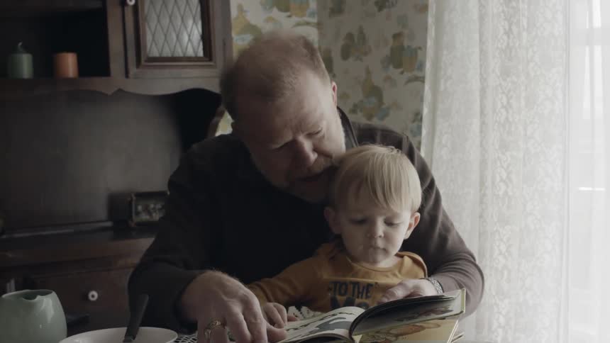 Grandfather is reading book to grandson at table