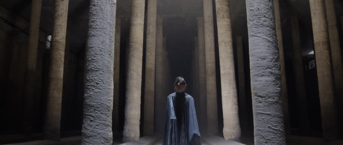 Man wearing a black cape dances in between rows of pillars