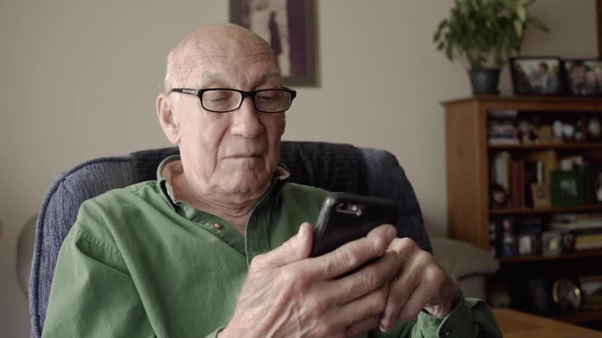 An old man is using a cellphone while sitting inside a living room in daylight