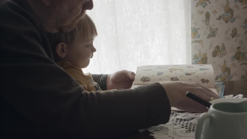 A man sits at a table holding his grandson and showing him books