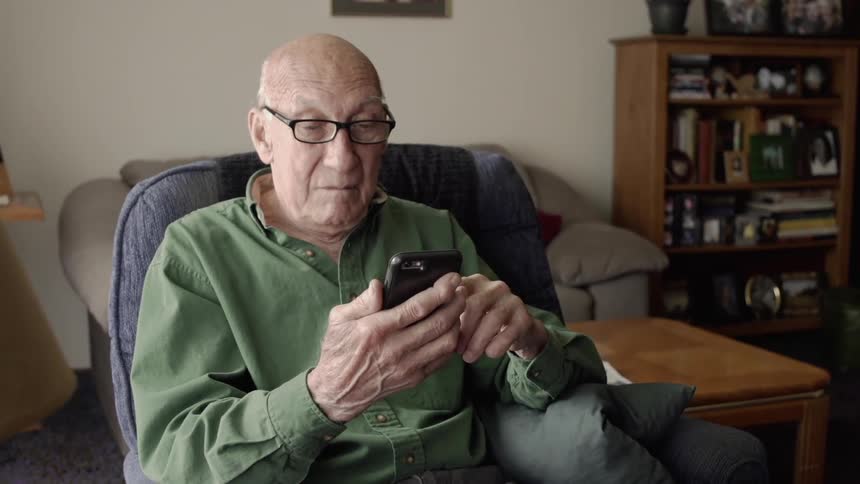 Older man sitting in chair in home is looking at cell phone
