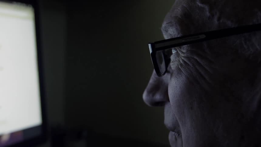 An older man is squinting as he looks at a computer screen in the dark